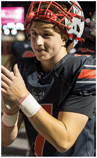A person wearing a football helmet and black jersey with red and white accents is looking to the side with a slight smile while standing outdoors at night. Descripci n generada con IA