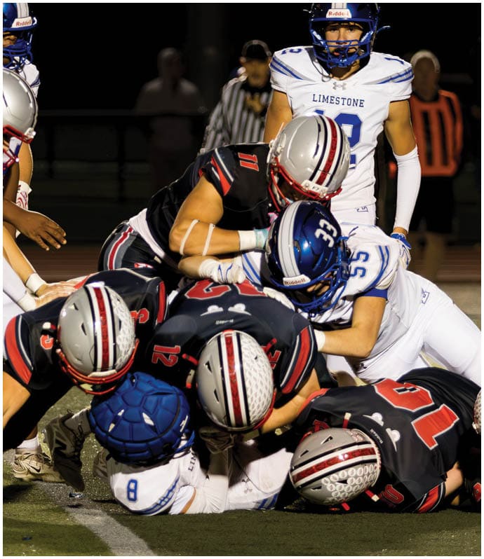 Several football players in black and white uniforms are engaged in a tackle on a grassy field during a night game. Descripci n generada con IA