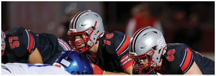 Two people in football uniforms and helmets are crouched in a ready position on a field facing an opponent in a white uniform. Descripci n generada con IA