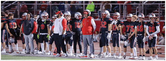 A group of people wearing football helmets and uniforms stand on the sidelines of a stadium filled with a large crowd of spectators. Descripci n generada con IA