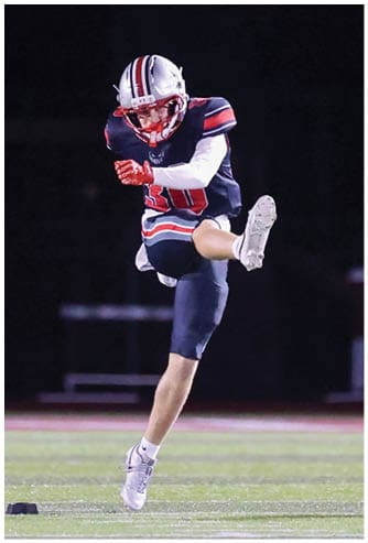A person wearing a white helmet and a dark sports jersey is looking down while a football is in mid-air against a dark background. Descripci n generada con IA