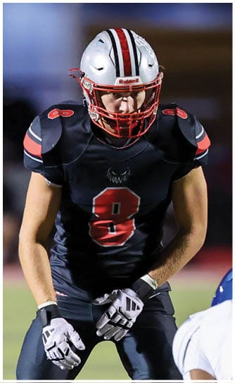 A person wearing a black football uniform with the number 8 and a white helmet is standing on a football field in a ready position. Descripci n generada con IA