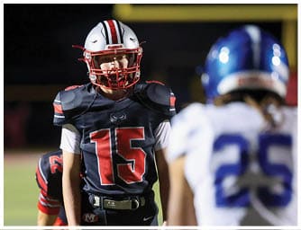 A person wearing a black and red football uniform with the number 15 stands on a field facing two people in white and blue football uniforms. Descripci n generada con IA