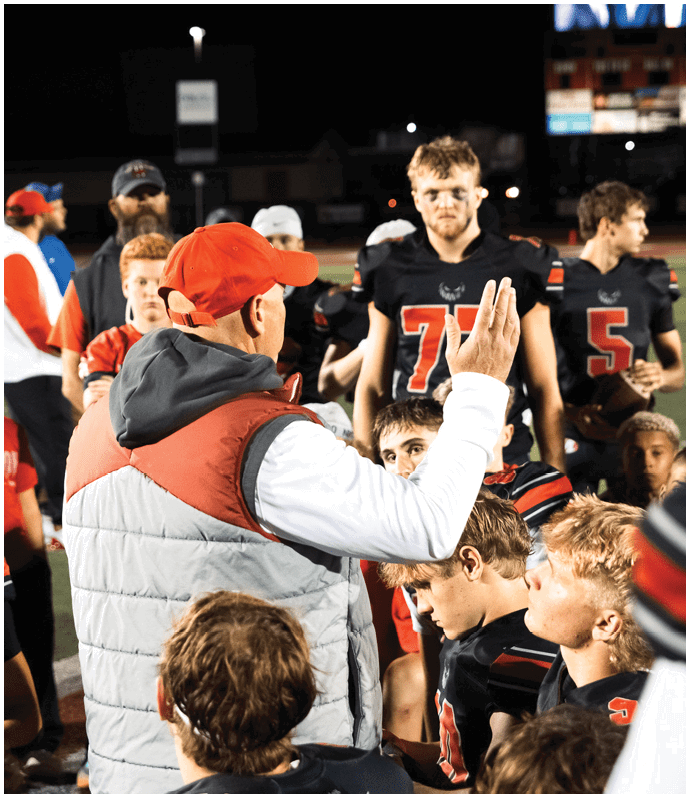 A person wearing a red cap and a gray and red vest is speaking to a group of people in black and red sports uniforms on a field at night. Descripci n generada con IA