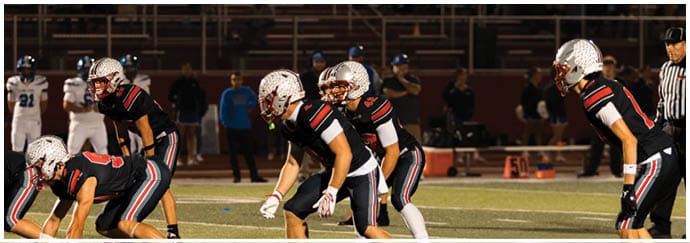 Several people wearing football helmets and uniforms are positioned on a field, appearing ready for a play during a nighttime game. Descripci n generada con IA