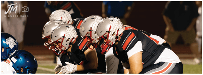 Several people in football uniforms and helmets are crouched in a line on a field facing opponents in blue helmets. Descripci n generada con IA