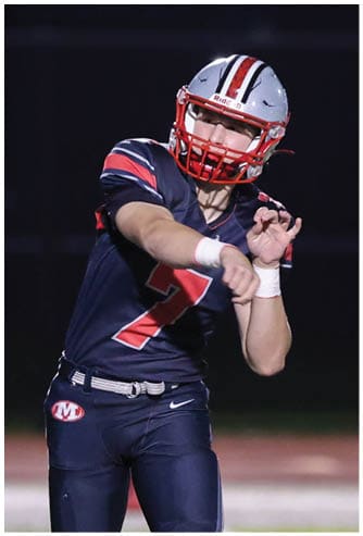 A person wearing a football uniform and helmet is throwing a football on a lit field at night while another person in a different uniform raises their arms nearby. Descripci n generada con IA