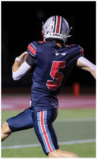 A person in a sports uniform is running on a football field while holding a football. Descripci n generada con IA