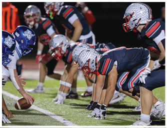 Three people in blue helmets and white uniforms are lined up in a football stance facing three people in black and red uniforms on a field. Descripci n generada con IA