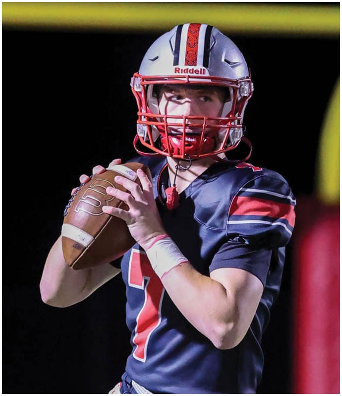 A person wearing a football helmet and uniform is holding a football in a throwing position on a field with a goalpost in the background. Descripci n generada con IA