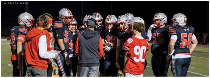 A group of football players and coaches wearing helmets and uniforms are gathered closely on a field at night, appearing to discuss strategy. Descripci n generada con IA
