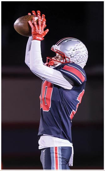 A person wearing a football uniform and helmet is catching a football with both hands raised above their head against a dark background. Descripci n generada con IA