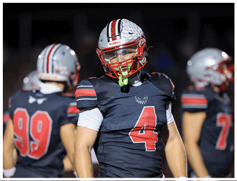 People wearing dark football uniforms with red and white accents stand on a field, with one person in the foreground wearing a helmet and a jersey numbered 4. Descripci n generada con IA