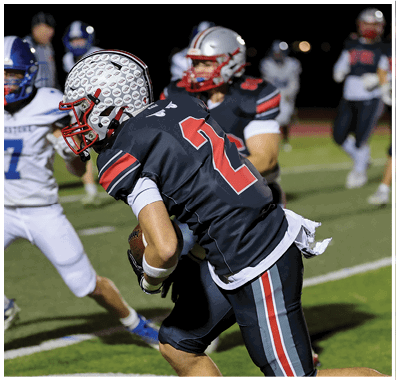 A group of people wearing football uniforms are actively playing on a field at night with one person in a black uniform running and others in white and black uniforms in the background. Descripci n generada con IA