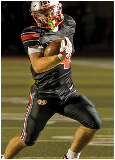 A person wearing a black football uniform and helmet is running on a football field while holding a football securely. Descripci n generada con IA