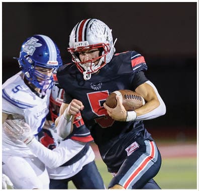 A person in a black football uniform is running with the ball while being chased by several people in white and blue football uniforms on a field. Descripci n generada con IA
