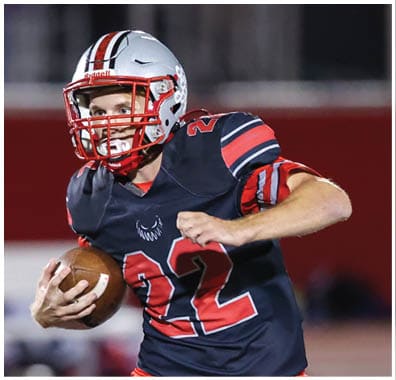 A person wearing a football uniform and helmet is holding a football and appears to be running or preparing to throw it on a field. Descripci n generada con IA