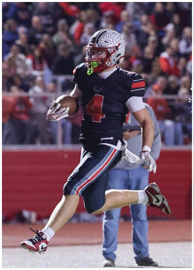 A person in a black football uniform with the number four is running with the ball while another person in a white uniform and blue helmet chases behind on a football field. Descripci n generada con IA