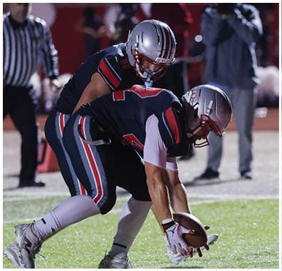 Two people wearing football helmets and uniforms are positioned closely on a field, with one person bent over and the other standing behind. Descripci n generada con IA