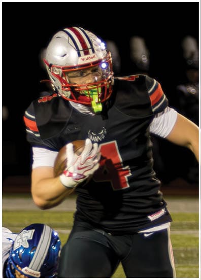 A person in a black football uniform is running with the ball while two people in white uniforms with blue helmets chase and one is on the ground on a football field at night. Descripci n generada con IA