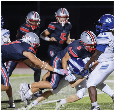 Several football players in dark uniforms and helmets are actively engaged in a play on a field under stadium lights. Descripci n generada con IA
