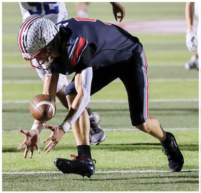 A person in a black football uniform is bending down to catch a football while another person in a similar uniform and two people in white uniforms are running behind on a football field at night. Descripci n generada con IA