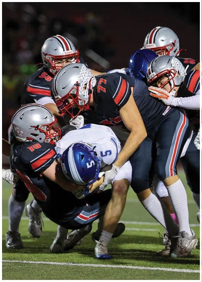 Several football players in dark uniforms are tackling a player in a white and blue uniform on a grassy field during a game. Descripci n generada con IA