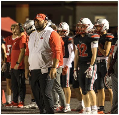 A group of people wearing football helmets and uniforms stand on the sidelines while a person in a white and red hoodie with a red cap and headset looks on during a nighttime football game. Descripci n generada con IA