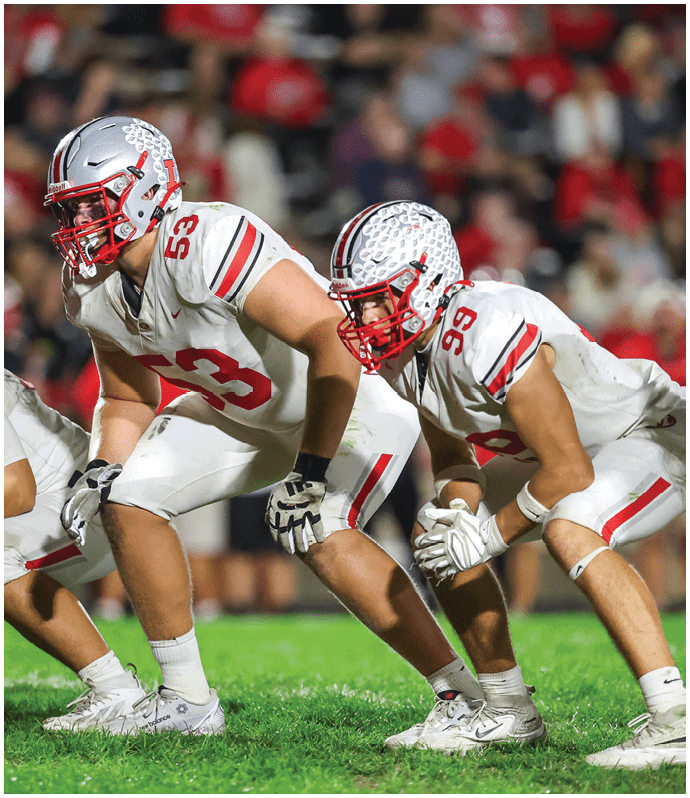 Three football players in white uniforms and helmets are crouched in a ready position on a grassy field during a game. Descripci n generada con IA