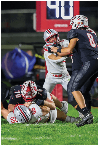 Players in football uniforms are actively engaged in a tackle on a grassy field during a game. Descripci n generada con IA