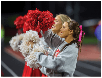 People wearing casual clothing and holding red and white pom-poms are cheering enthusiastically at an outdoor event, likely a sports game. Descripci n generada con IA