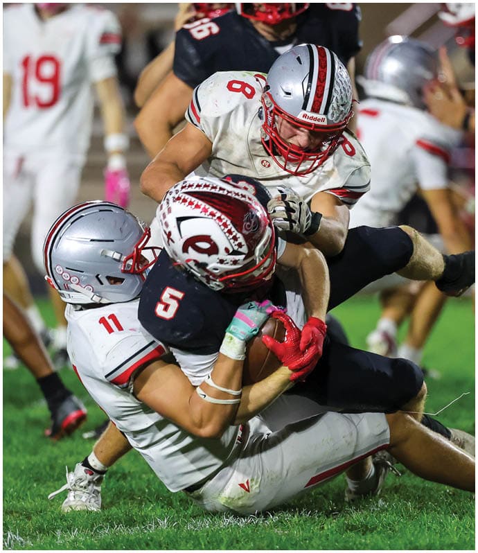 Three football players in helmets and uniforms are engaged in a tackle on a grassy field during a game. Descripci n generada con IA