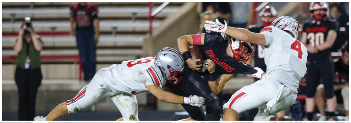 Two football players are engaged in a tackle on the field with blurred spectators and a photographer in the background. Descripci n generada con IA