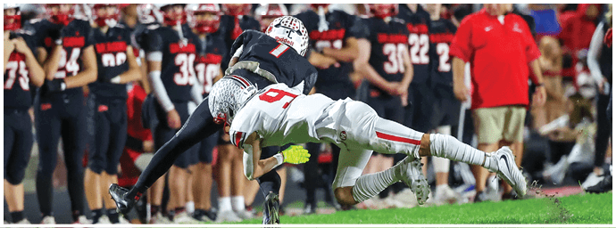 Two people in football uniforms are engaged in a tackle on a grassy field with others standing in the background. Descripci n generada con IA