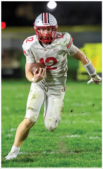 A person wearing a white and red football uniform with the number 12 is running on a grassy field while holding a football. Descripci n generada con IA