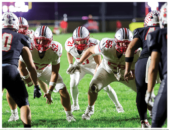 Several people in football uniforms and helmets are positioned on a grassy field, appearing ready for a play during a nighttime game. Descripci n generada con IA