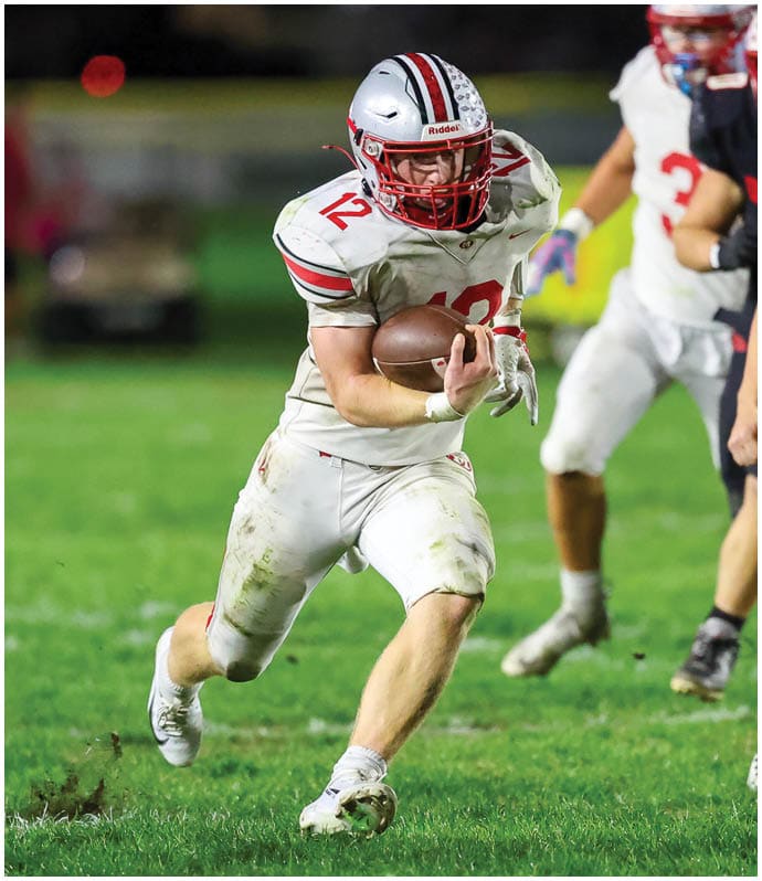 A person in a white football uniform with the number 12 is running on a grassy field while holding a football, with other players in the background. Descripci n generada con IA