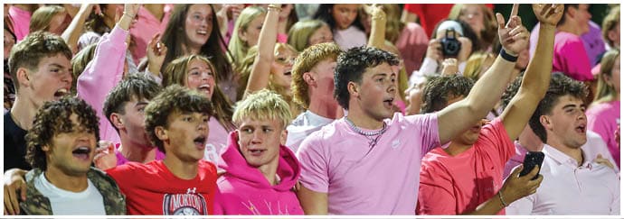 A group of people wearing pink shirts enthusiastically cheer and raise their hands outdoors, with some holding cameras. Descripci n generada con IA