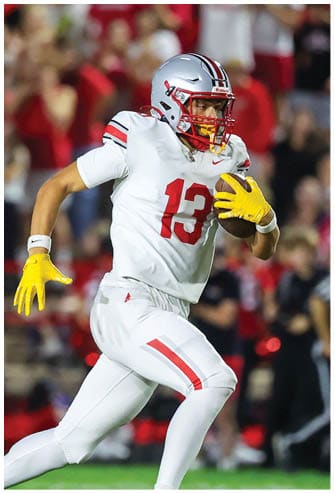 A person wearing a white football uniform with the number 13 and yellow gloves is running while holding a football. Descripci n generada con IA