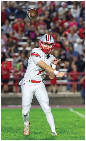 A person wearing a white football helmet with red stripes and a red faceguard is partially visible on the left side with a blurred crowd in the background. Descripci n generada con IA