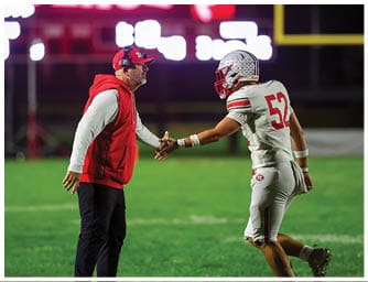 A person wearing a red and white hoodie and cap is shaking hands with a football player in white and red uniform on a green field at night. Descripci n generada con IA