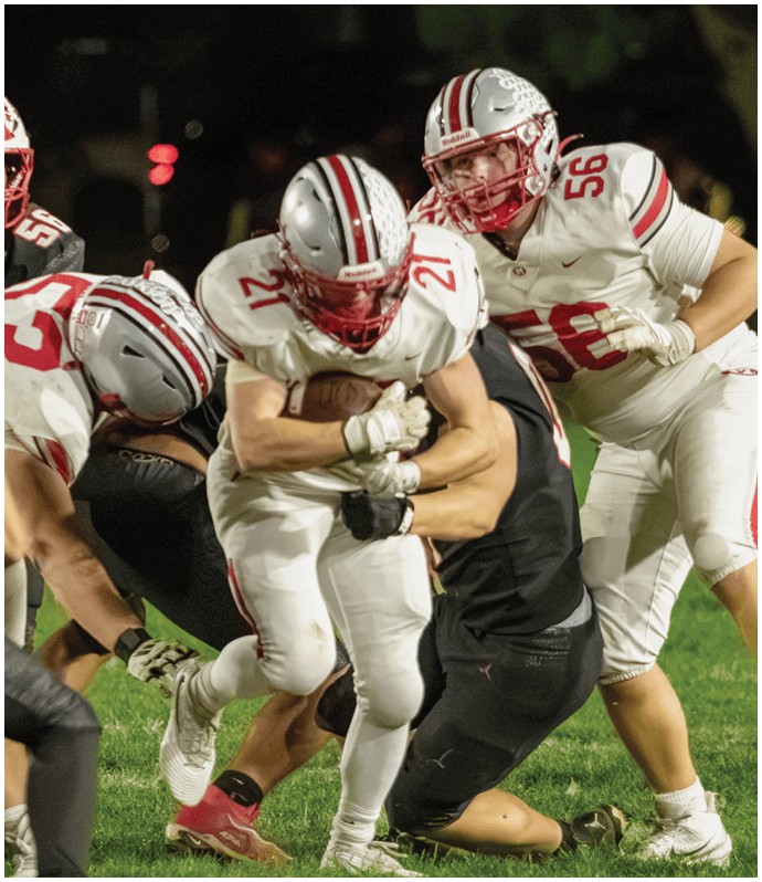 Two people wearing football helmets and uniforms are engaged in a play, with one person holding the football tightly while the other appears to be supporting or protecting them. Descripci n generada con IA