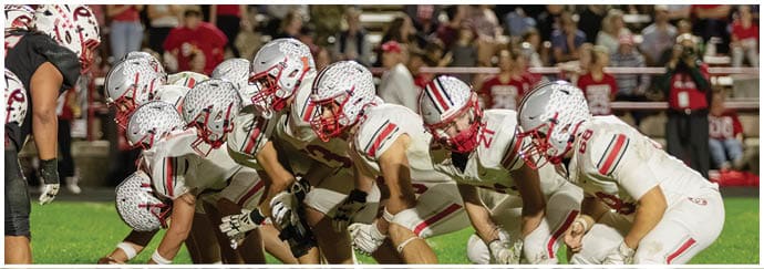 A group of football players in white uniforms and helmets are lined up on the field in a ready stance during a game with spectators in the background. Descripci n generada con IA