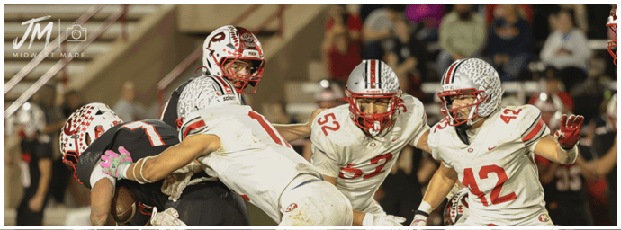 Several people in football uniforms are engaged in a play on a field, with some players tackling and others preparing to block or catch. Descripci n generada con IA