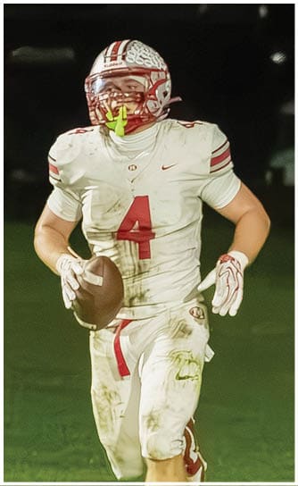 A person wearing a white football uniform with the number four and a helmet with a brain design is standing on a field at night. Descripci n generada con IA