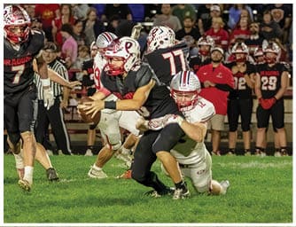 A person in a black football uniform is being tackled by a person in a white football uniform while other players and spectators watch in the background. Descripci n generada con IA