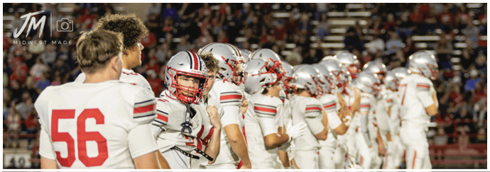 A line of people wearing white sports uniforms with red and black stripes stand on a grassy field, likely preparing for a football game. Descripci n generada con IA