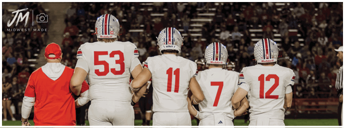 Four people in football uniforms and helmets walk arm in arm on a field with a crowd in the background. Descripci n generada con IA