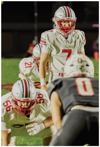 People in football uniforms are positioned on a field, preparing for a play during a nighttime game. Descripci n generada con IA