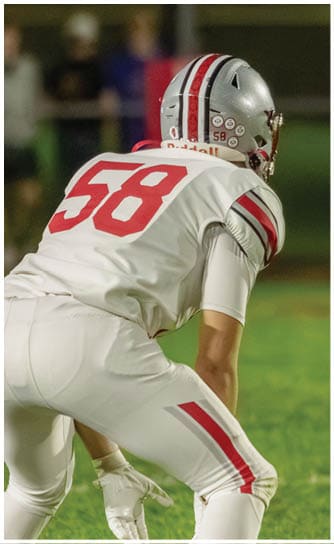A person wearing a silver football helmet with red stripes is on a field with a large digital scoreboard displaying the number 25 in the background. Descripci n generada con IA